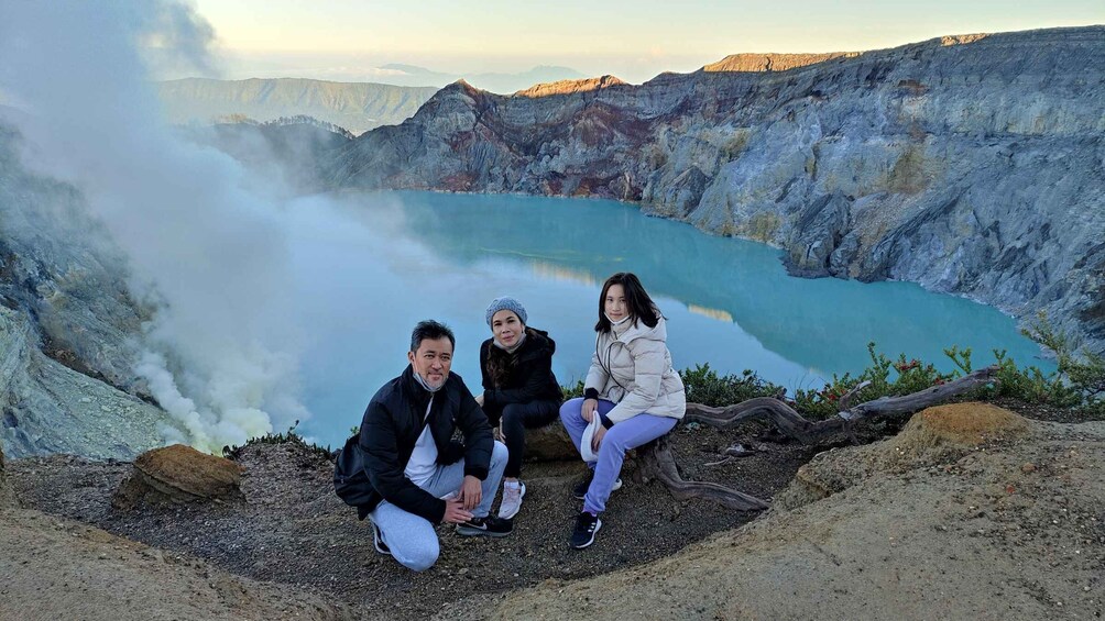 Three people sitting on a rocky outcrop overlooking a turquoise lake with steam rising from the ground. 2