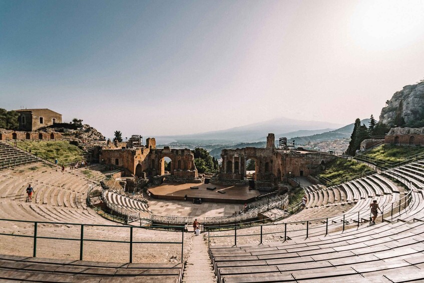 An ancient amphitheater with ruins and a stage, surrounded by hills and a distant cityscape. 4