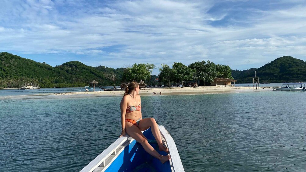A woman in a bikini sitting on a boat in a tropical bay with clear water, a sandy beach, and lush green hills in the background. 3