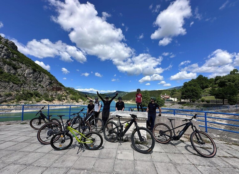 A group of cyclists posing on a dock with bicycles, a lake, and mountains in the background. 5