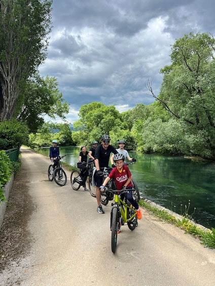 A group of cyclists on a paved path alongside a river, surrounded by lush greenery. 1