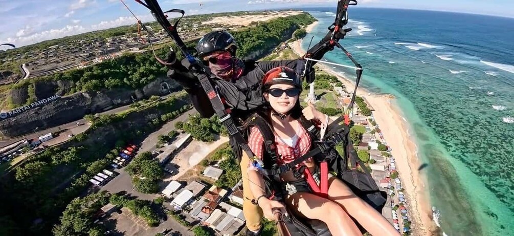 A person skydiving with a parachute over a coastal area with a beach and a large rock formation labeled 'Pantai Pandawa.' 2