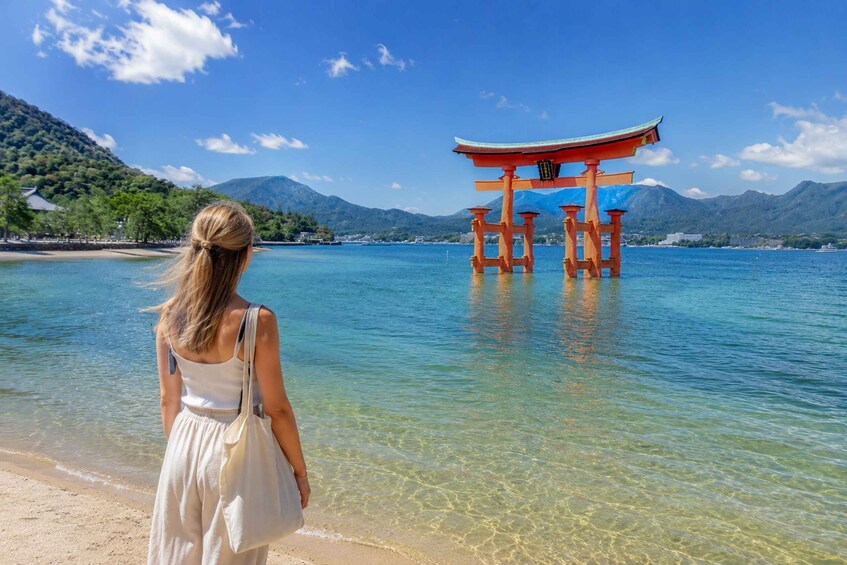 A woman walking on a beach with a traditional Japanese torii gate in the water. 1