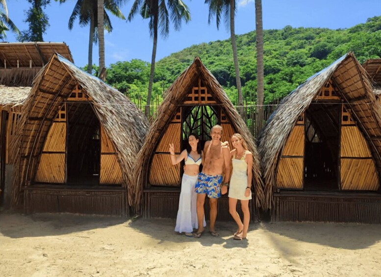 A group of people posing in front of thatched-roof huts on a sandy beach with palm trees in the background. 2