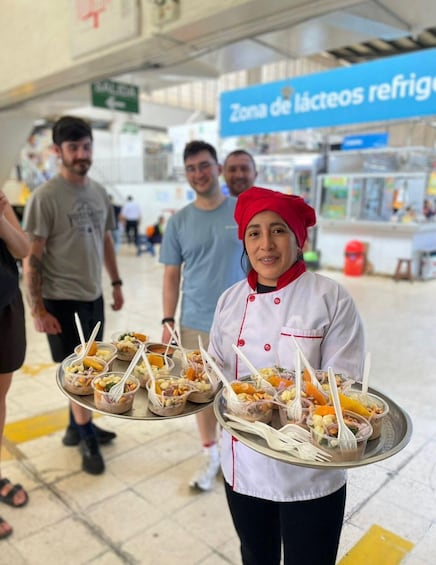 A person holding trays of food in a public area with a sign reading 'Zona de lácteos refrigerados' in the background. 1