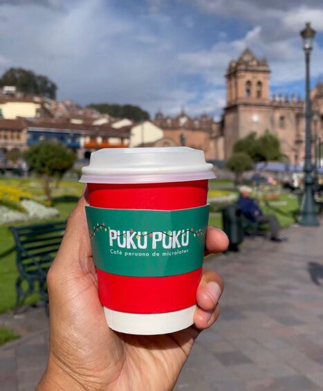A hand holding a red and white coffee cup with 'PUKU PUKU' branding, set against a blurred urban park background with a historic building. 3
