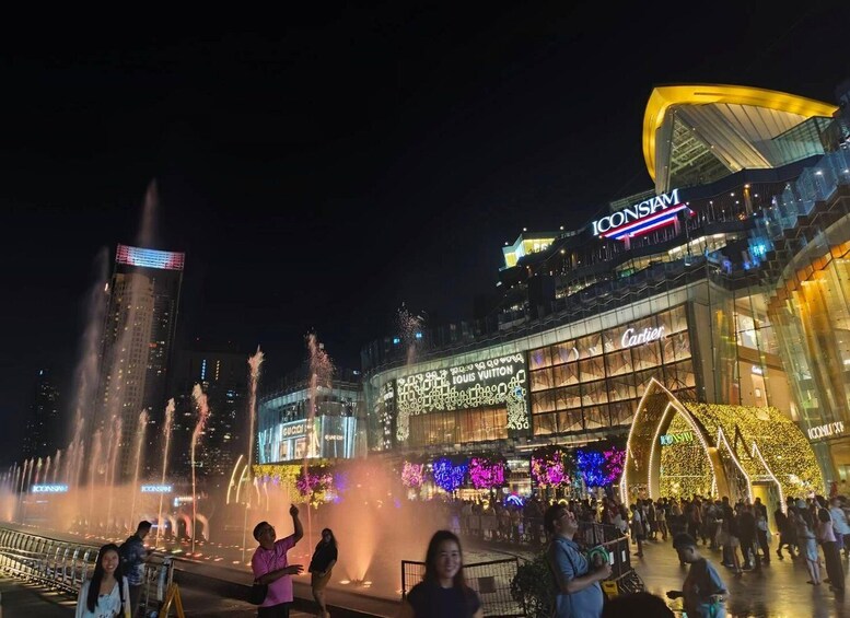 A night scene with a water fountain show, illuminated storefronts, and a crowd of people. 2
