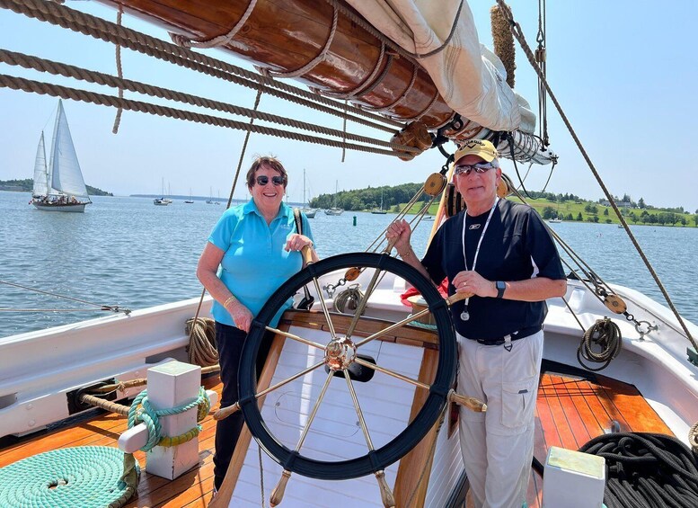 Two people on a sailboat with a wooden wheel, ropes, and a calm lake with other boats in the background. 5