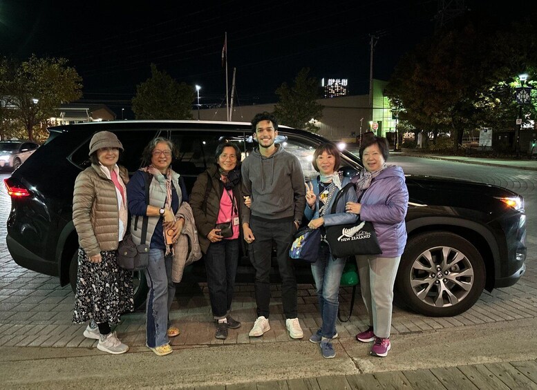 A group of people standing in front of a black SUV on a paved area at night. 4