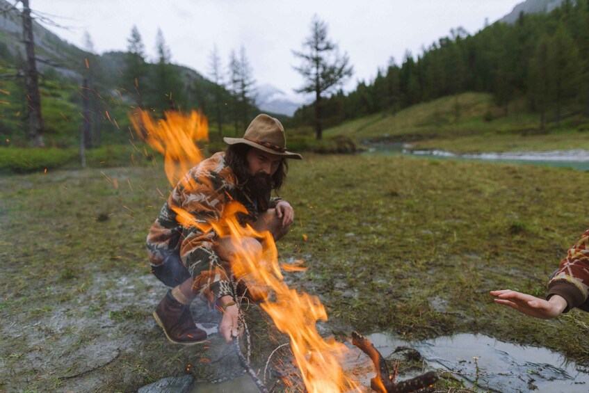 A person crouching near a campfire in a grassy field with a river and mountains in the background. 3