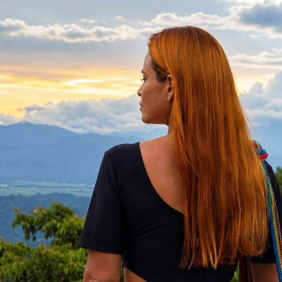 A person with long red hair wearing a black top, standing with a mountainous landscape in the background. 5