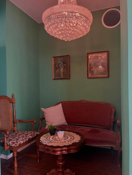 A room with a red sofa, a wooden chair, a table with a lace tablecloth, and two framed pictures on the wall. 3