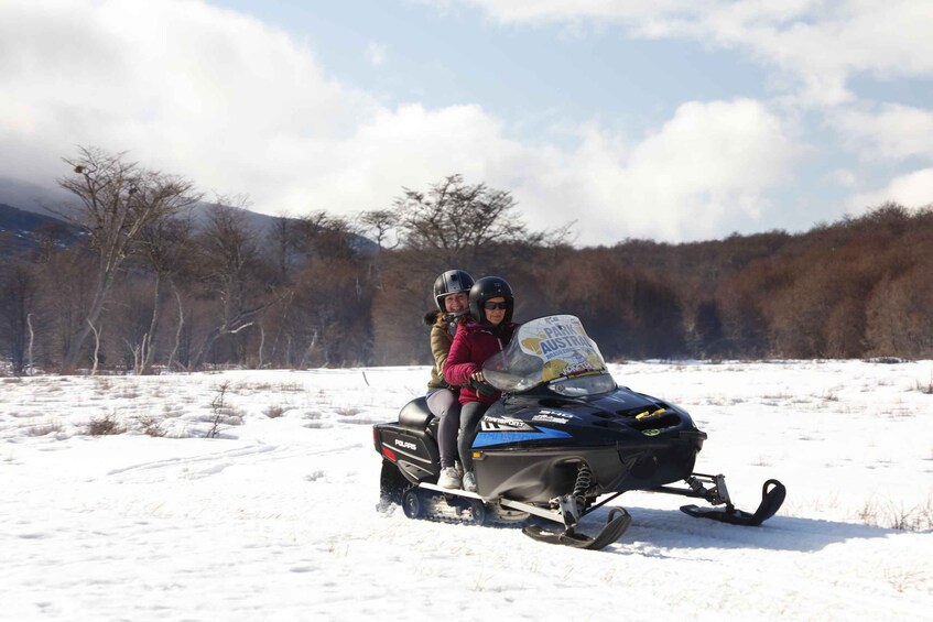 Two people on a snowmobile in a snowy field with trees and mountains in the background. 5