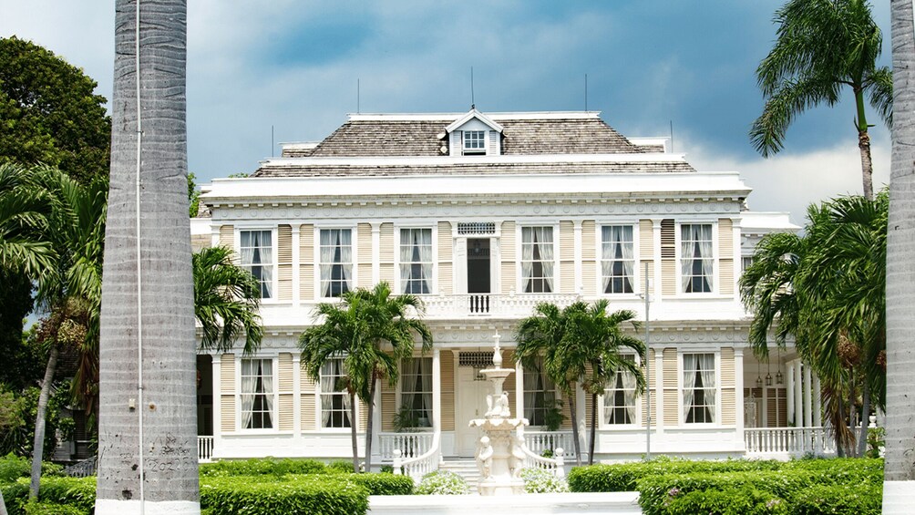 A large, white colonial-style building with a symmetrical facade, a central entrance with a decorative fountain, and palm trees surrounding the property. 2