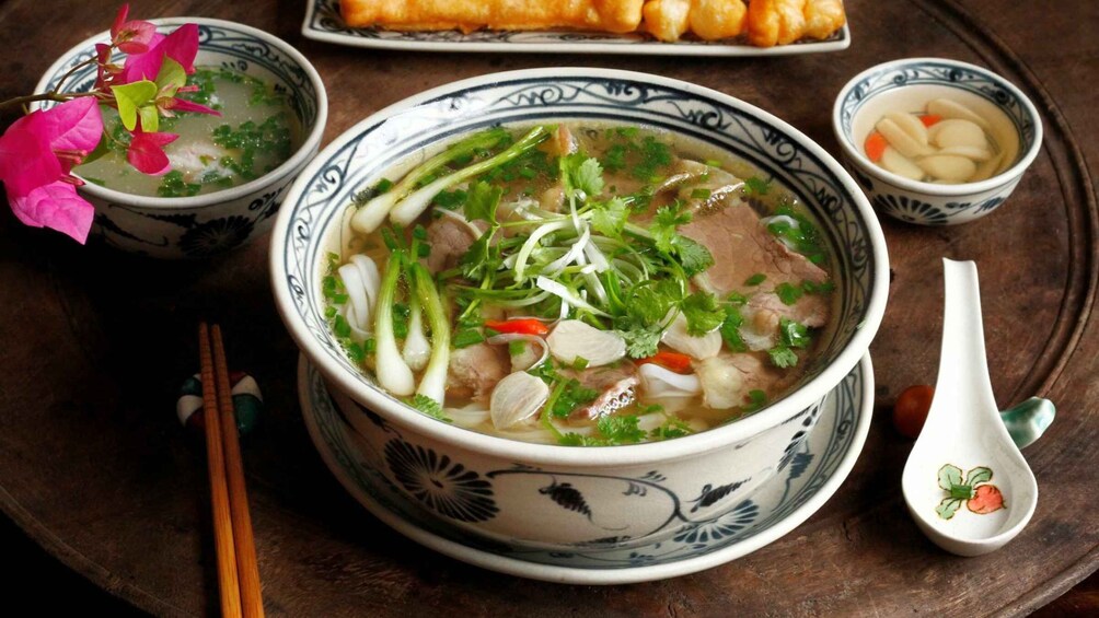 A bowl of soup with beef, vegetables, and herbs, accompanied by a plate of spring rolls and a small bowl of rice. 4