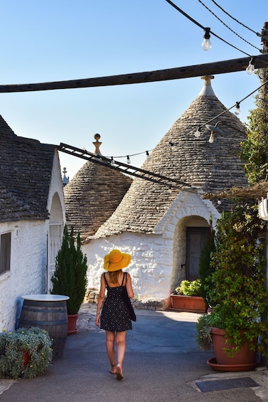 A woman in a black dress and yellow hat walking down a street with stone buildings and potted plants. 5