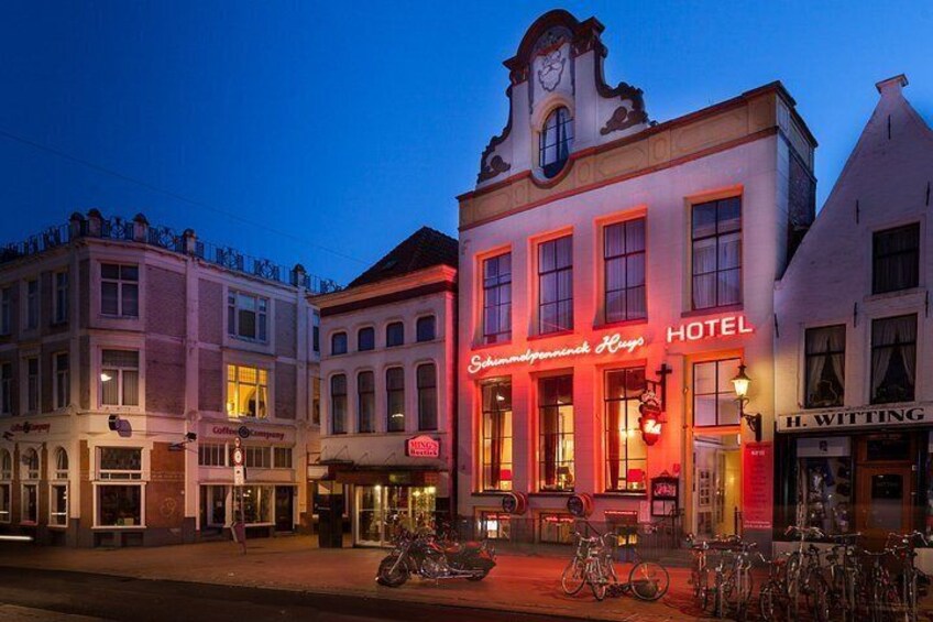 A hotel with a clock tower, bicycles parked outside, and other buildings with illuminated signs. 4
