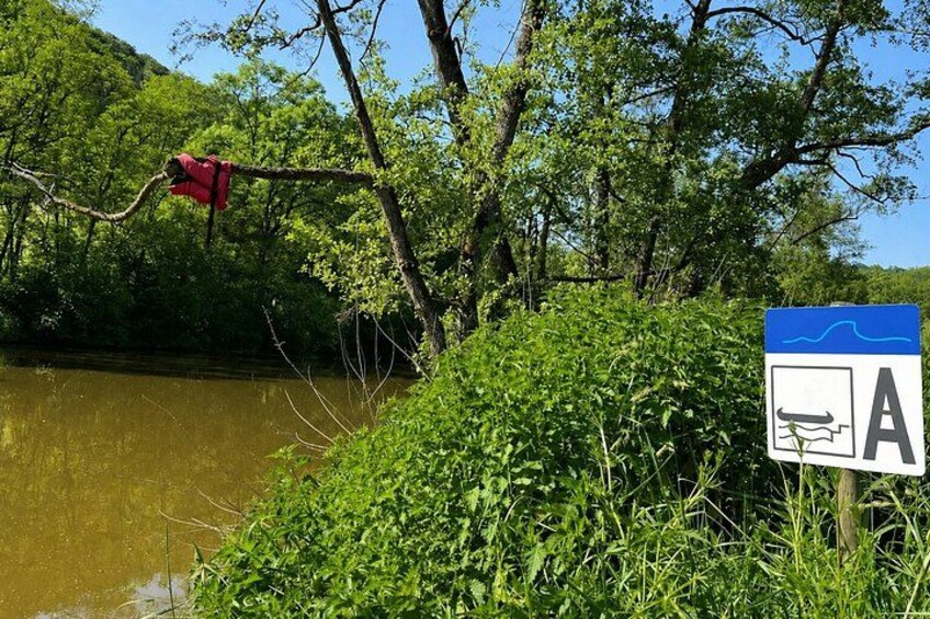 A riverbank with dense greenery, a blue sign with a white 'A' and a white boat symbol, and a red object hanging from a tree branch. 2