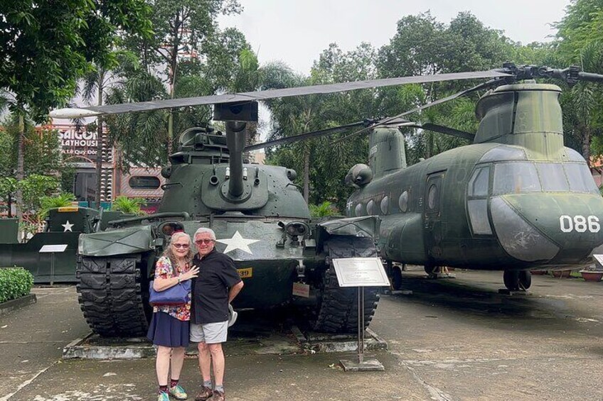 A military helicopter with the number 086, a couple standing in front of it, and a signboard in the background. 3