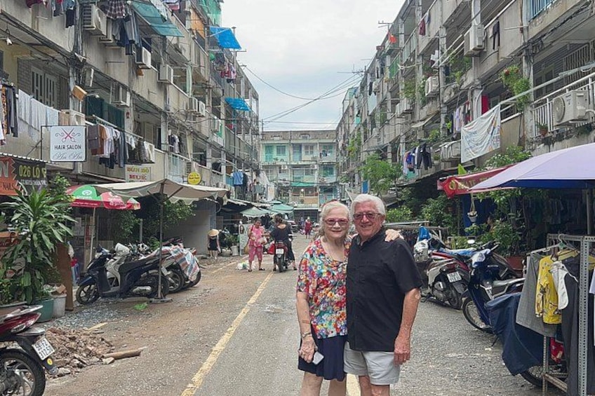 A narrow street lined with multi-story buildings, some with balconies and hanging laundry. There are parked motorcycles and a few pedestrians. 4