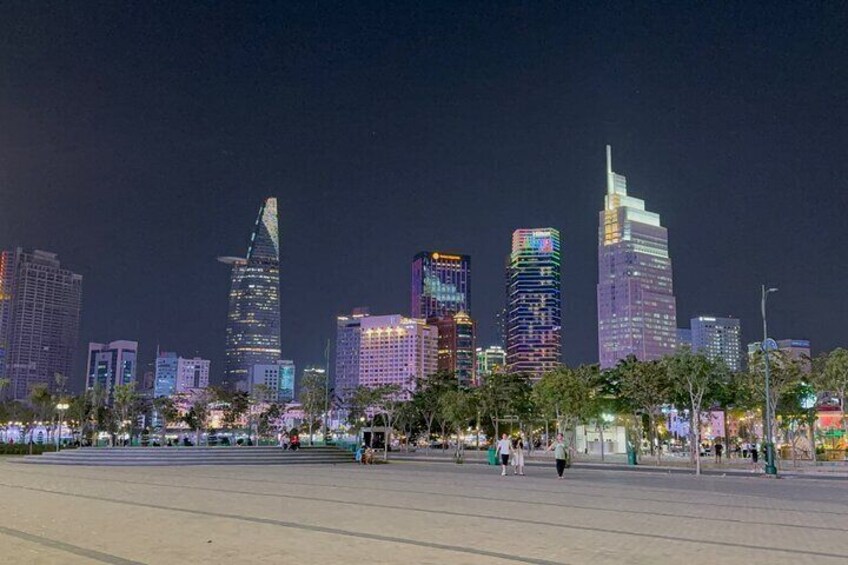 A city skyline at night with illuminated buildings and a wide pedestrian area. 1