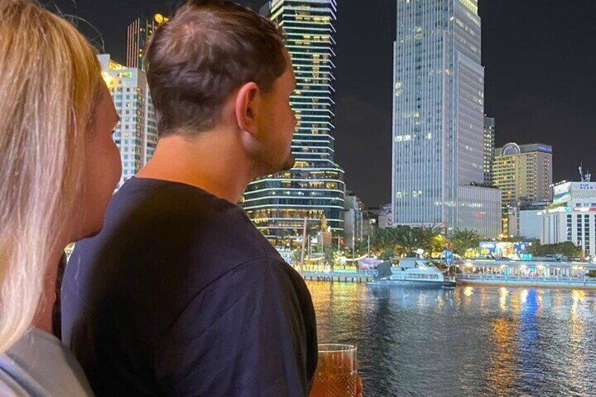 A couple overlooking a city skyline at night with illuminated buildings and a body of water. 2