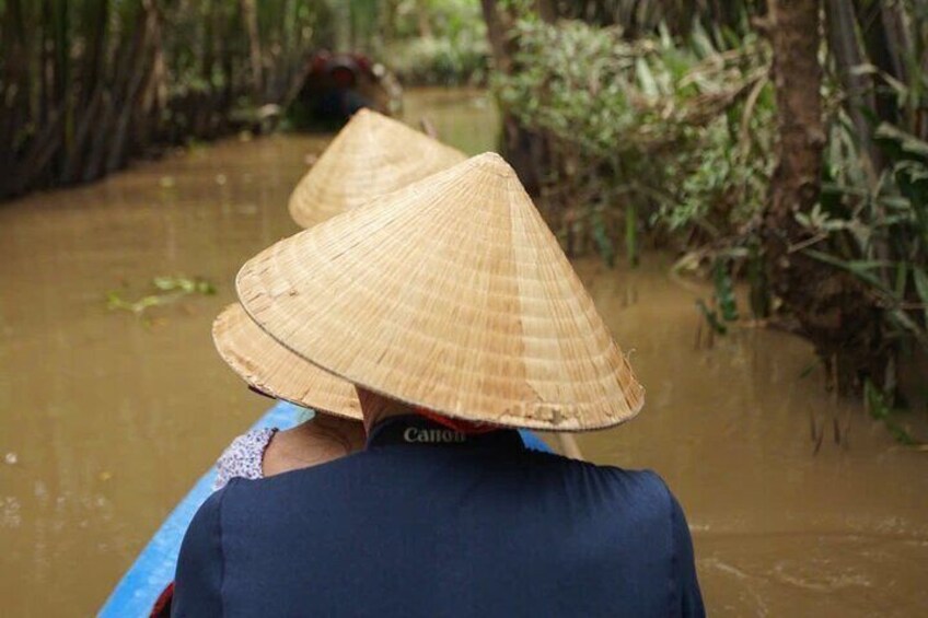 Two people in a boat wearing traditional conical hats, navigating a river surrounded by dense vegetation. 1
