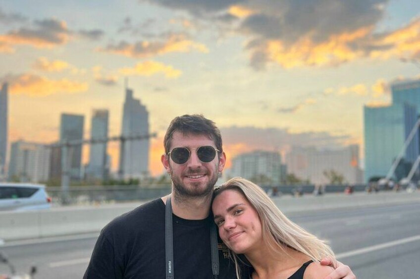 A couple standing in front of a city skyline with modern buildings and a sunset in the background. 5