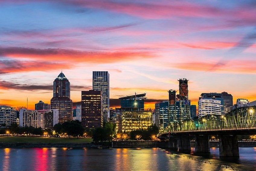 A city skyline at dusk with illuminated buildings and a bridge over a body of water. 1