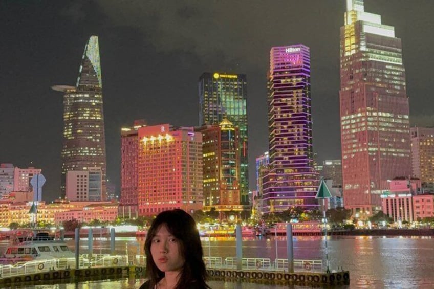 A nighttime cityscape with illuminated skyscrapers and a body of water in the foreground. 1