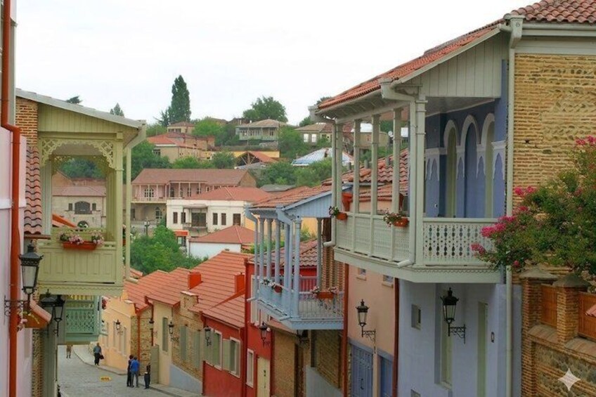A narrow street with colorful buildings, balconies with flowers, and a clear sky. 1