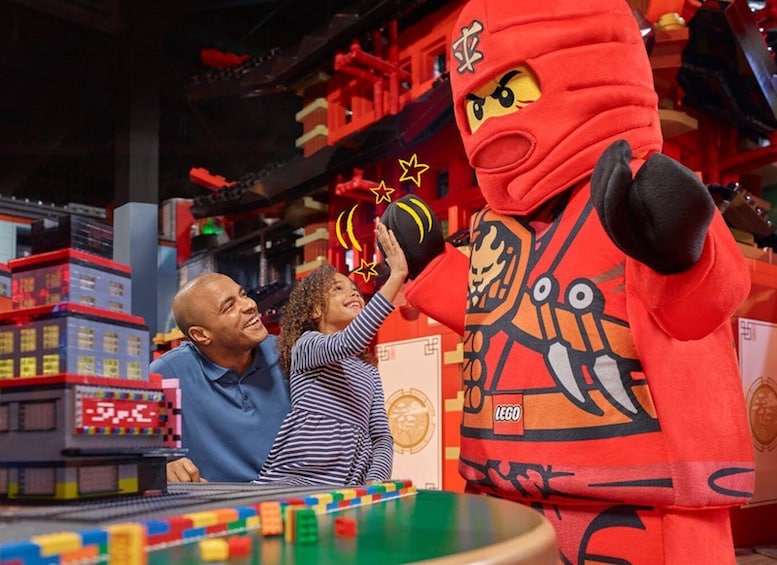 A LEGO figure interacting with a child and an adult in a LEGO-themed environment. 1
