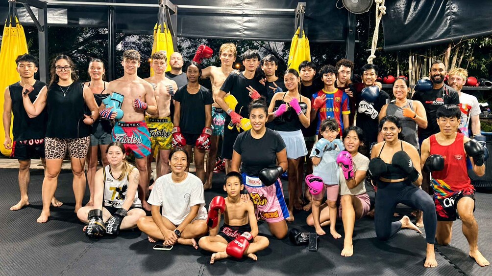 A group of people in a gym, some wearing boxing gloves and others in athletic wear, with a punching bag and gym equipment in the background. 1