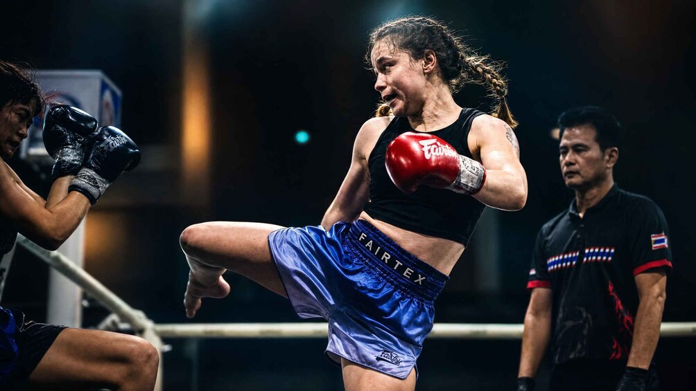 A female athlete in a black top and blue shorts with 'FAIRTEK' on them, performing a high kick in a boxing ring. 2