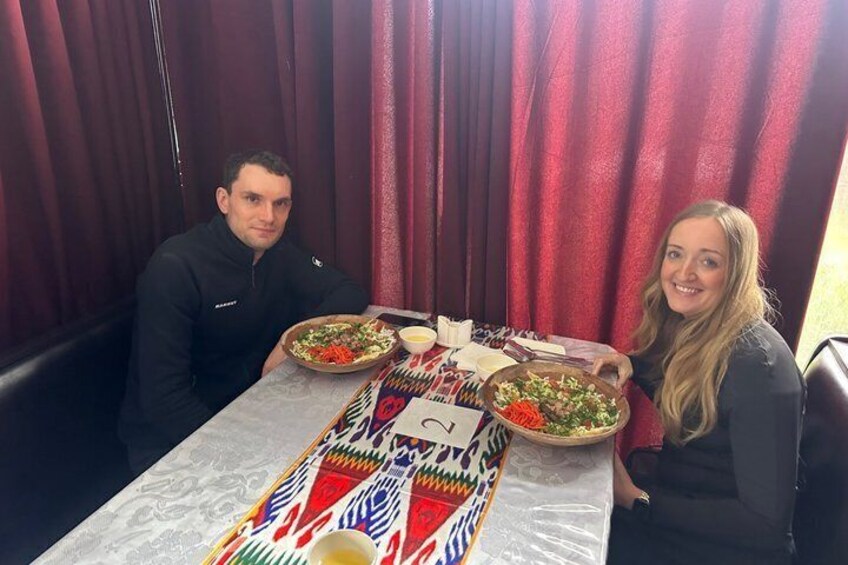 Two people seated at a table with a patterned tablecloth, plates of food, and a glass of drink. 3