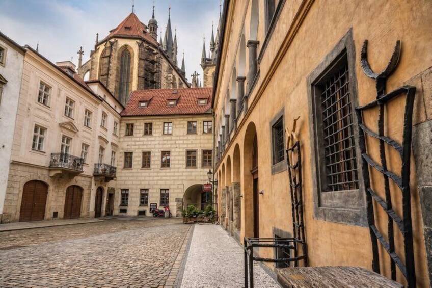 A cobblestone street with historic buildings, a church with a red roof, and a wrought iron gate. 3