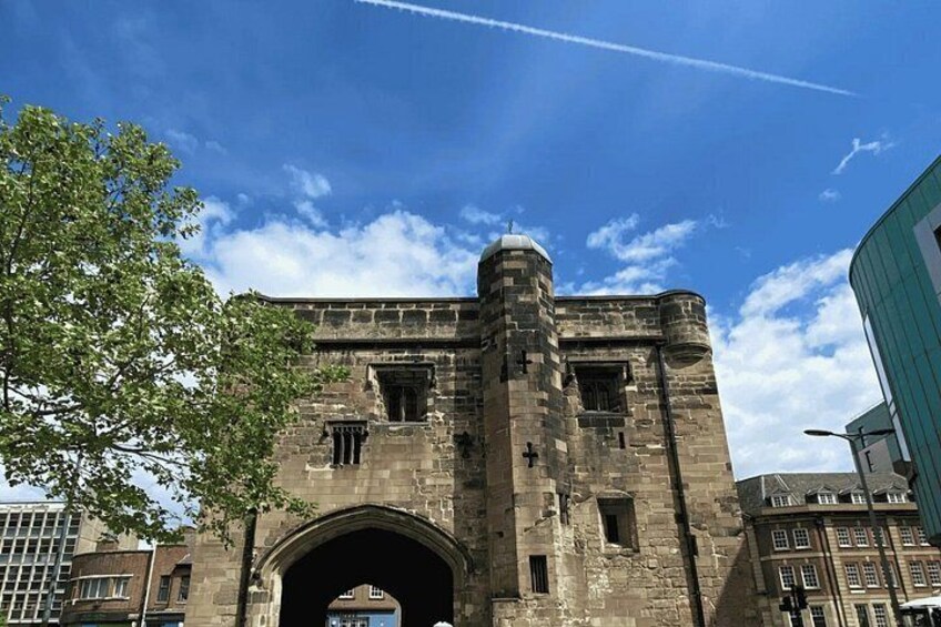 A historic stone building with a large archway, a turret, and a modern glass structure nearby. 3