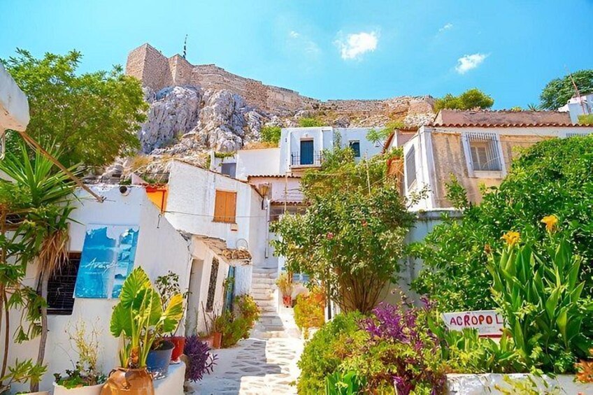 A narrow street with white buildings, a sign reading 'ACROPOL,' and a mountain in the background. 1