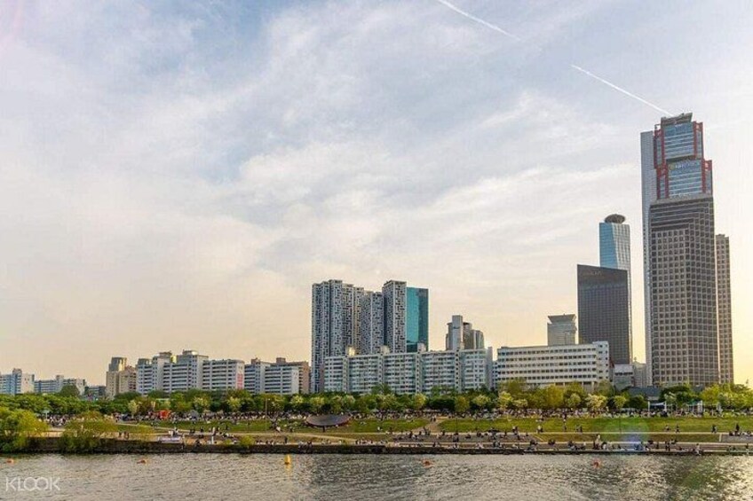A city skyline with modern high-rise buildings, a river in the foreground, and a park with people. 5