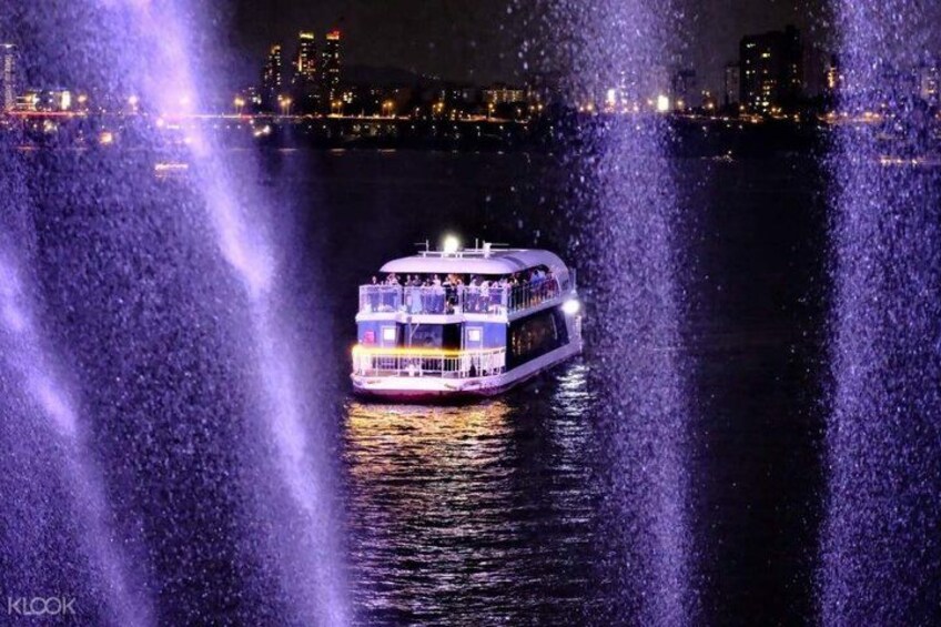 A brightly lit riverboat with multiple decks, surrounded by water fountains, and a city skyline in the background. 3