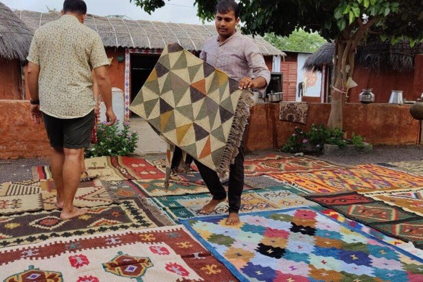 Two people carrying a large, patterned fabric outdoors, with traditional buildings and a thatched roof in the background. 5