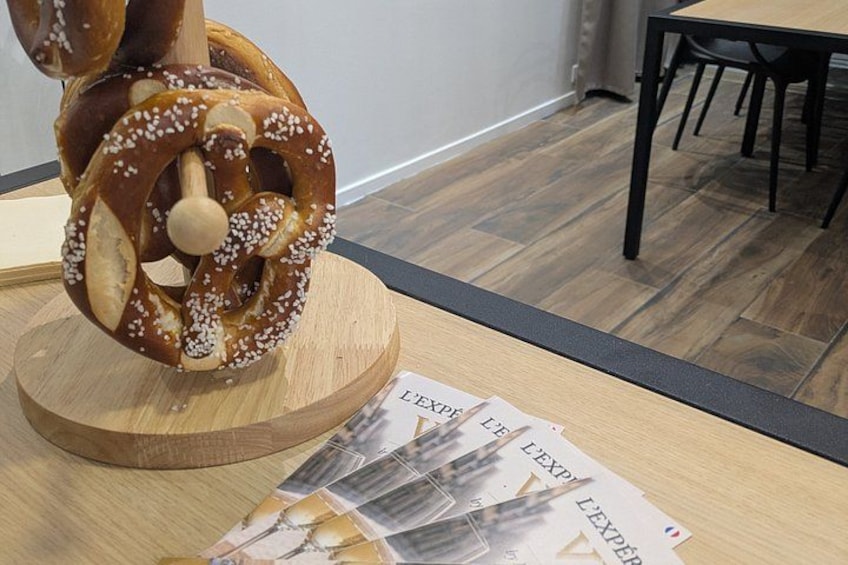 A pretzel on a wooden stand, a table with papers, and a dining area with chairs and a table. 1