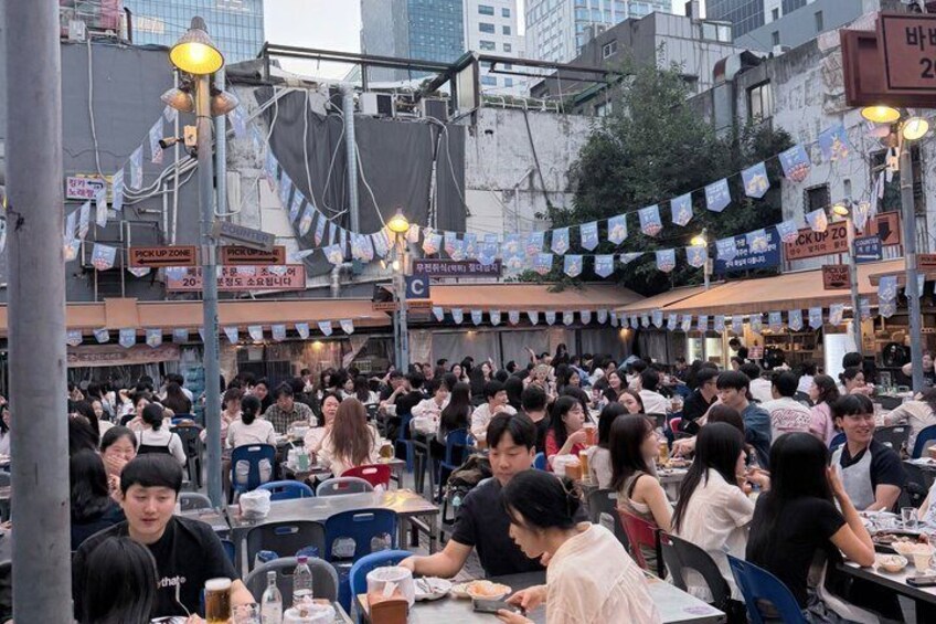 A crowded outdoor dining area with tables and chairs, surrounded by buildings and street signs. 4