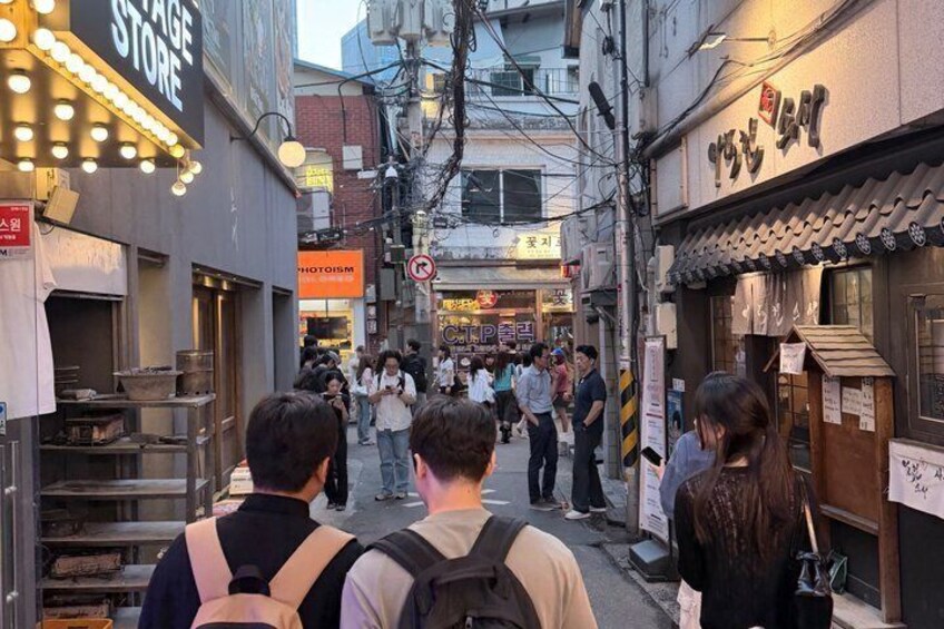 A narrow street with people walking, a food stall, and a building with a sign. 3