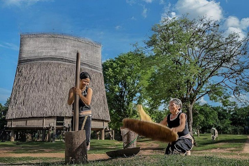 Two people near a traditional thatched structure, a large tree, and a grassy area. 2