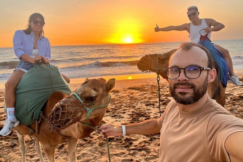 A beach scene with a camel and two people riding it, a sunset in the background, and a person taking a selfie. 1