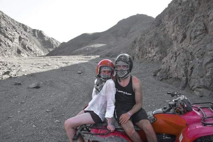 Two people on a red ATV in a rocky terrain with mountains in the background. 1