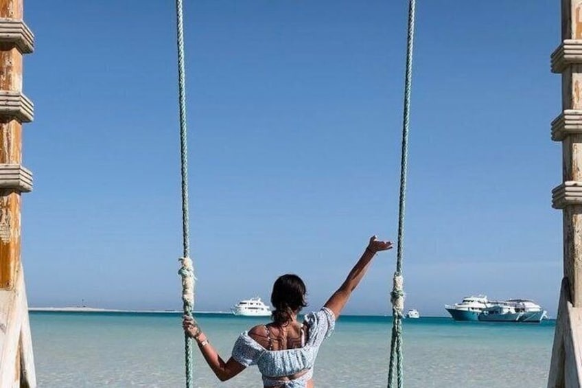 A person on a swing with ropes, a clear blue sky, and a boat in the background. 5