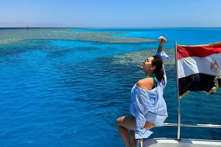 A person on a boat with a flag, clear blue water, and a distant shoreline. 1