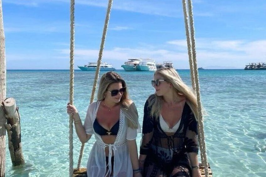 Two people on a swing over clear turquoise water with boats in the background. 2